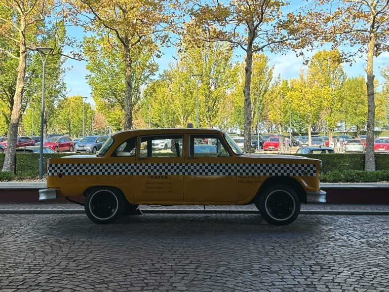 Yellow taxi parked near trees and cars in a sunny outdoor setting.
