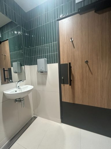 Interior of a restroom with a sink, wooden stall door, and green tiled walls.