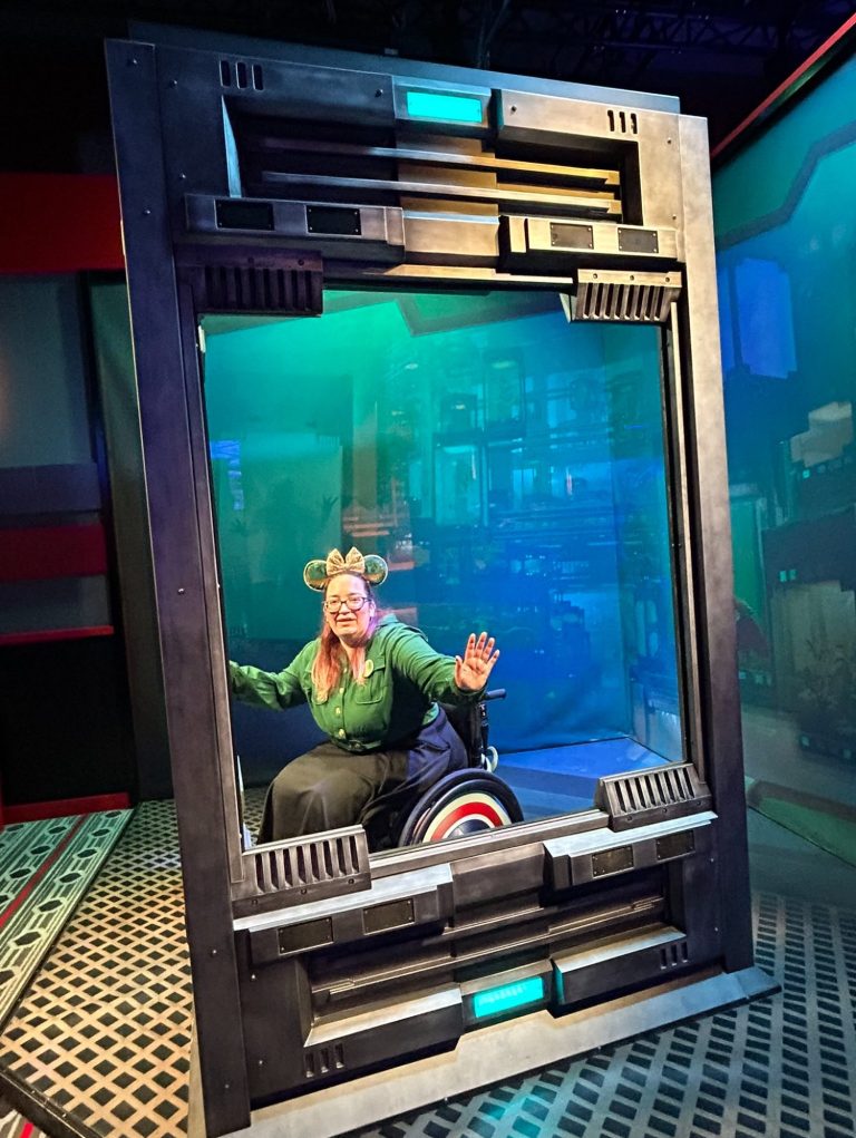 A person in a futuristic glass chamber, smiling and waving, with blue and green lighting.