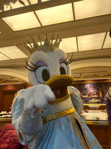 Daisy Duck in a blue gown and crown, waving cheerfully.