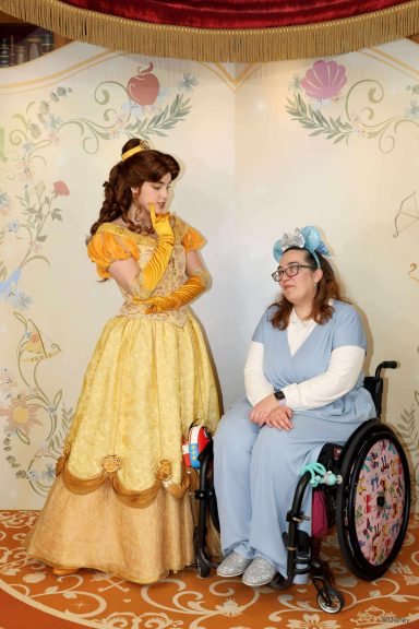 A young woman in a wheelchair with a blue flower crown sits beside a character in a yellow gown.