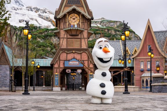 Olaf the snowman stands in front of a quaint alpine building with snow-capped mountains.