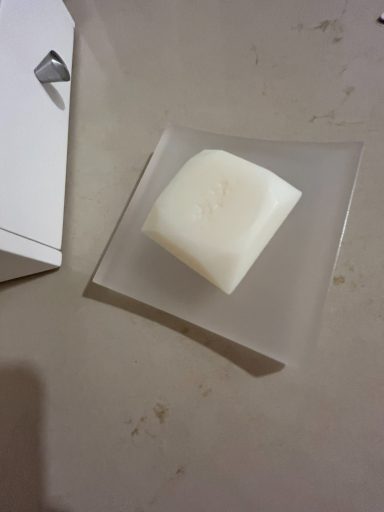 White soap bar on a clear dish, resting on a light surface next to a white box.