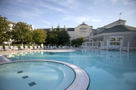 Outdoor pool with a jacuzzi, surrounded by trees and a hotel in the background.