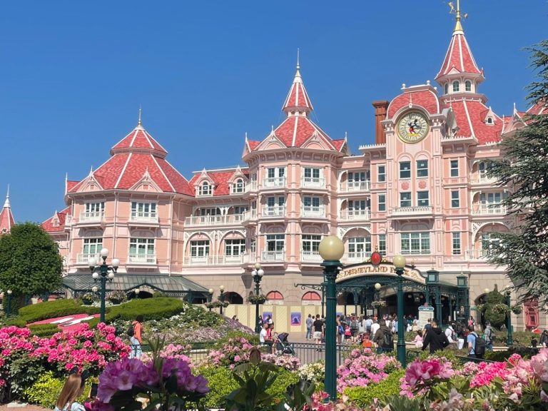 Pink castle-like building with turrets, surrounded by flowers and visitors in a sunny setting.
