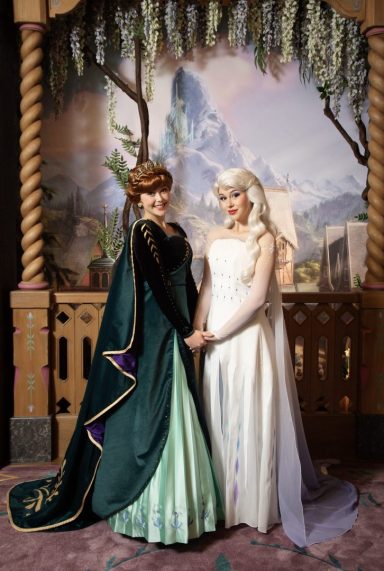 Anna and Elsa stand together, surrounded by a scenic backdrop.