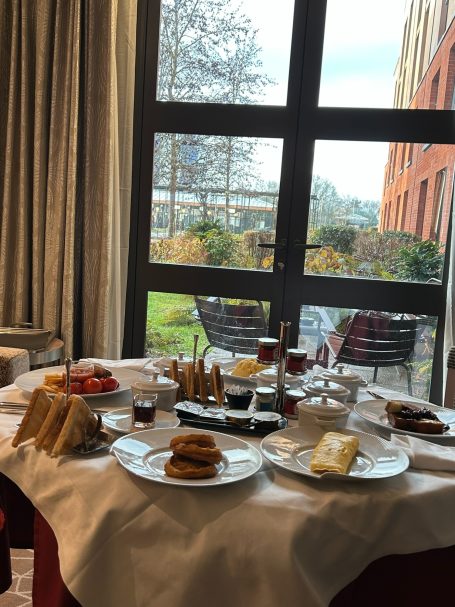 A dining table set with various dishes and drinks, with a view of greenery outside.