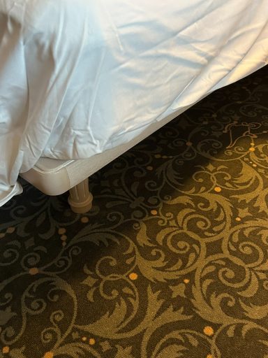 Bed with a white coverlet on patterned carpet, showing part of the bed frame.