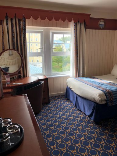 A hotel room with a bed, desk, mirror, and window showing greenery outside.