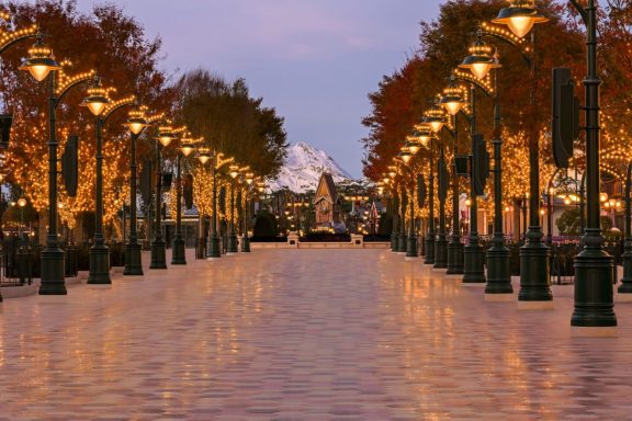 A lit walkway lined with trees, leading towards a distant mountain at dusk.