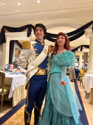 A prince and princess in royal attire pose together in a regal setting.