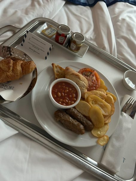A tray with breakfast including sausages, baked beans, croissant, and condiments.