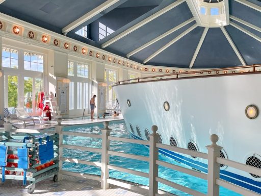 Indoor pool with a boat-like structure and lounging areas. Sunlight streams through large windows.