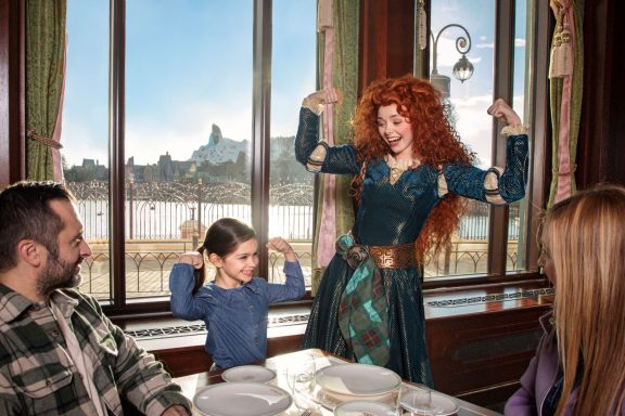 Merida poses playfully with a young girl in a restaurant with a scenic view.